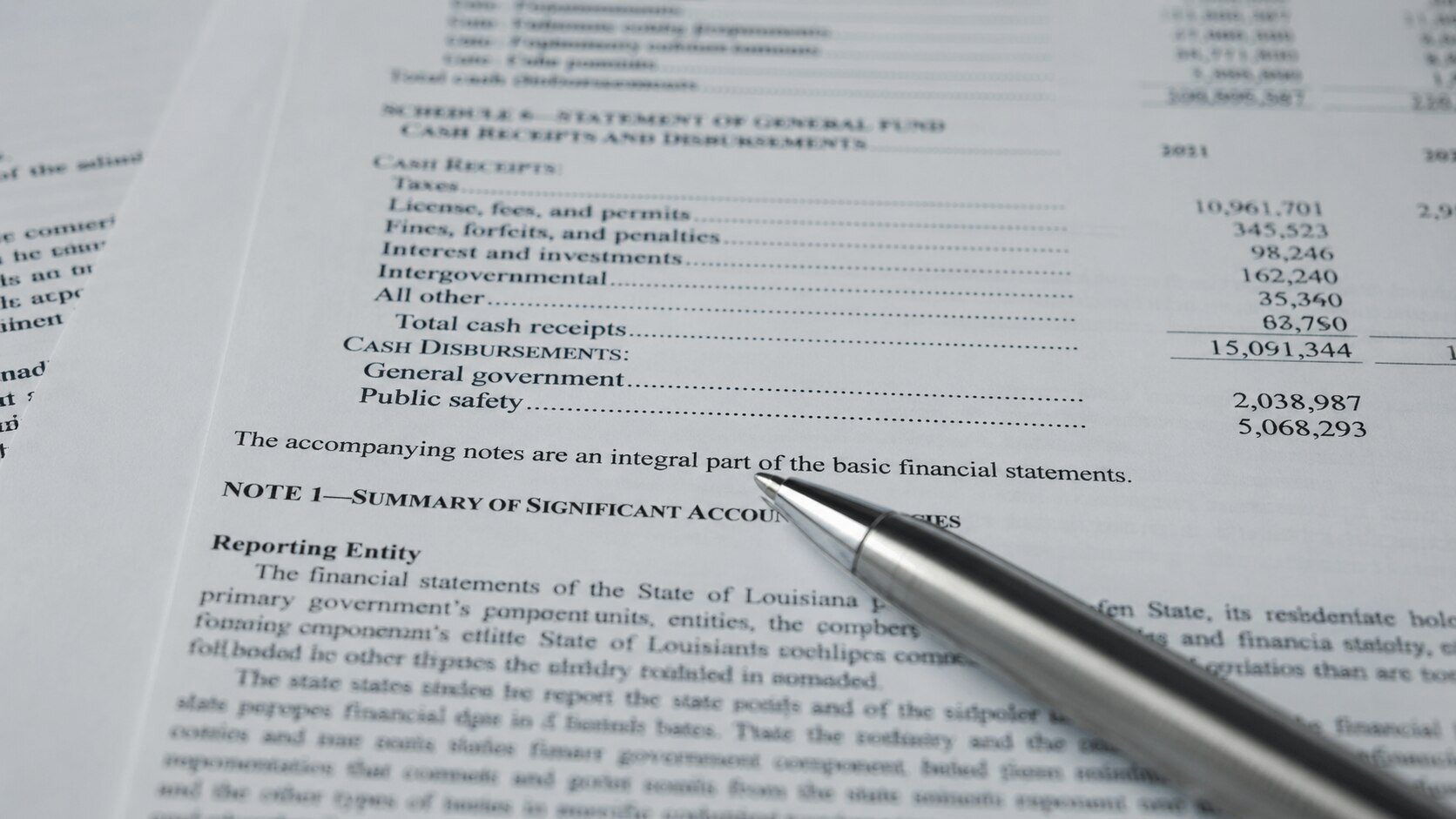 Close-up of a government financial statement showing cash receipts, disbursements, and accounting notes, with a silver ballpoint pen resting across the page — illustrating the kind of official document footnote in which the BEA disclosed its removal of investment silver from GDP trade data.