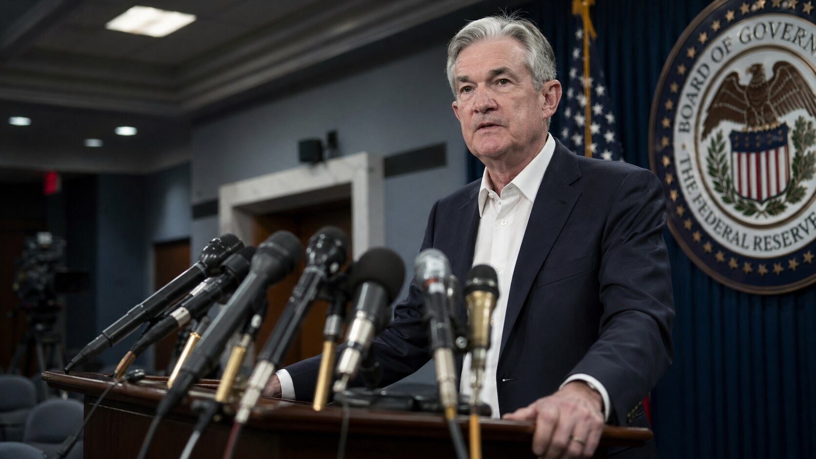Federal Reserve Chair Jerome Powell speaking at a press conference podium with multiple microphones, Federal Reserve seal visible in the background