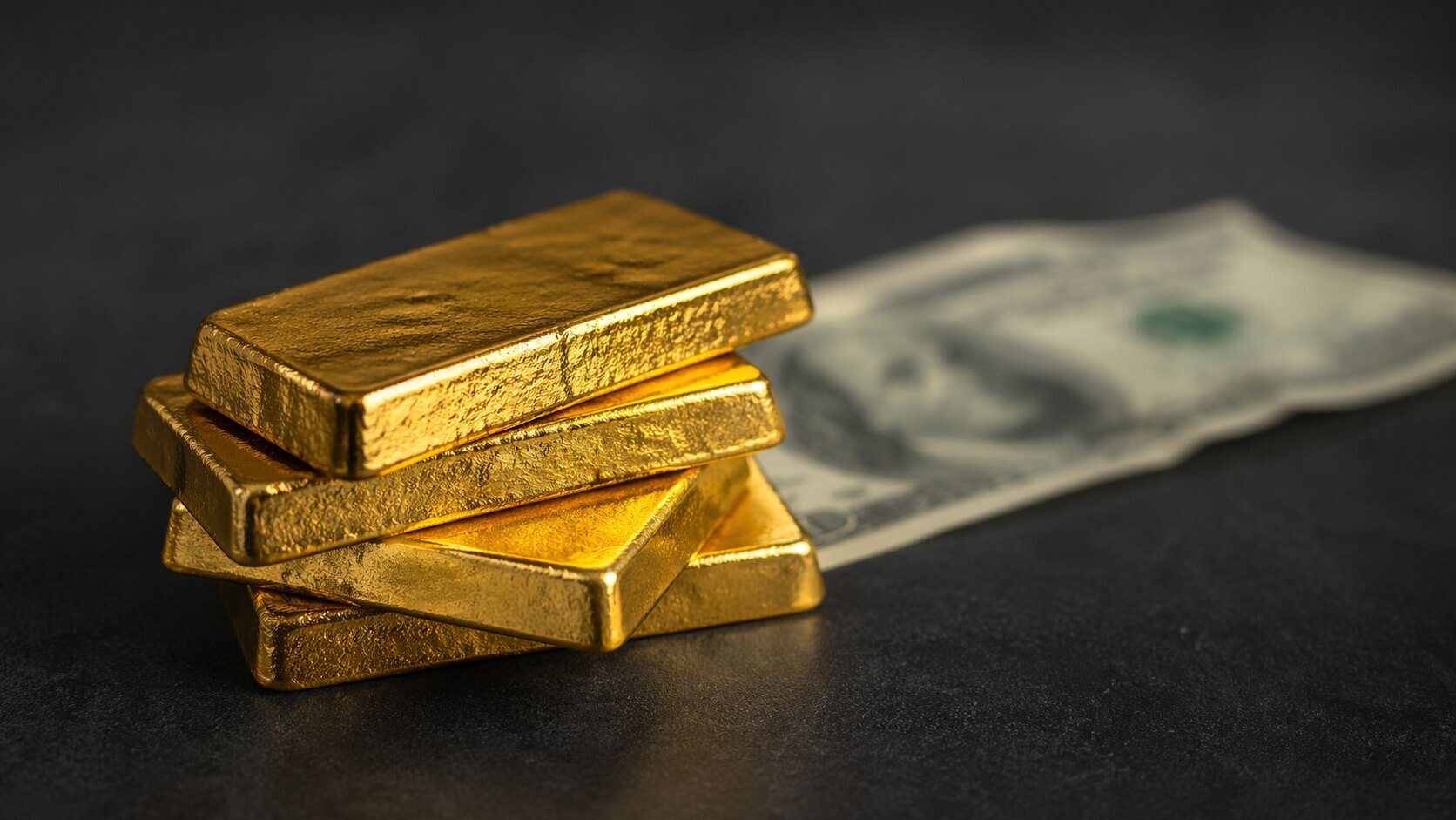 Gold bullion bars stacked on a dark surface with a U.S. hundred-dollar bill fading out of focus in the background